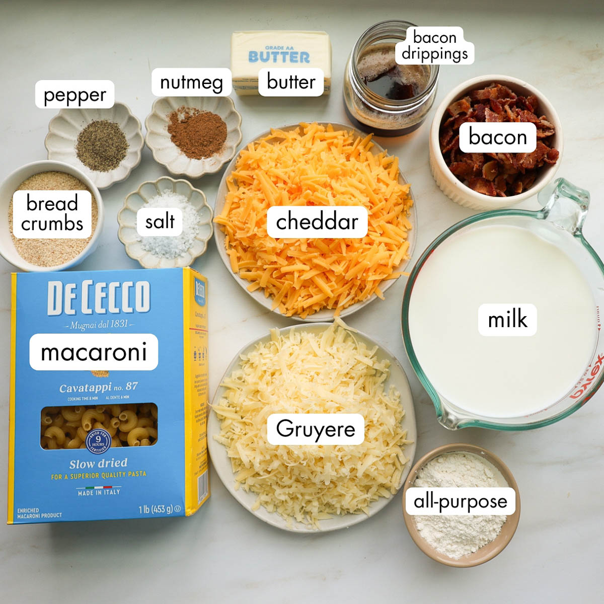All the ingredients for the baked macaroni and cheese, including milk, cheddar, Gruyere, bacon, bacon, drippings, butter, nutmeg, pepper, salt, bread crumbs, macaroni, and all-purpose flour.