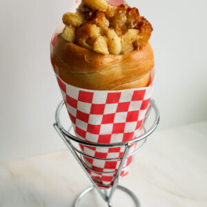 A bread cone wrapped in red and white checked paper and filled with bacon mac and cheese.