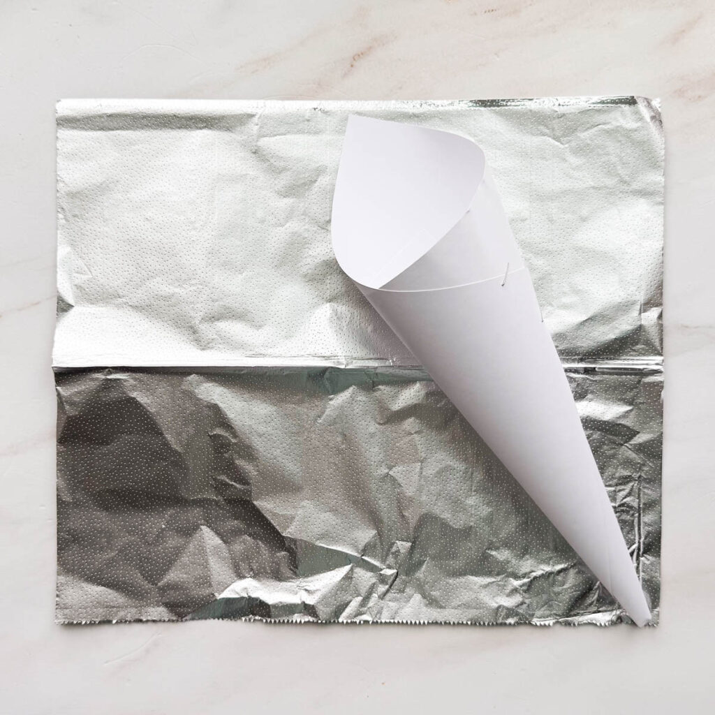 A white piece of paper rolled up in a cone shape, laying on top of a sheet of aluminum foil.