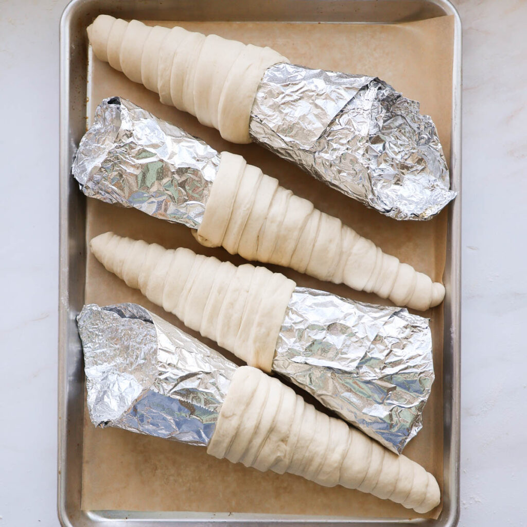 Four aluminum foil cone molds with bread dough wrapped around them, on a baking sheet lined with parchment paper.