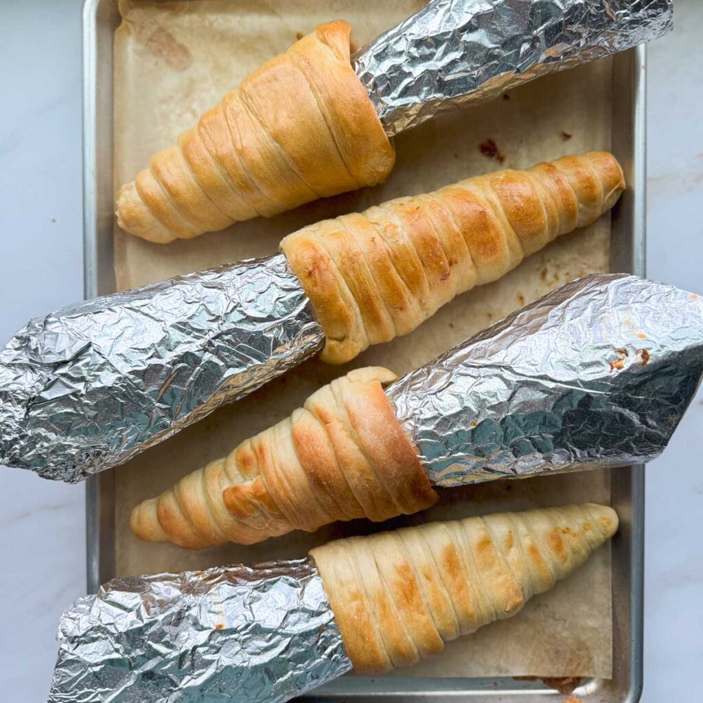 Four aluminum foil cone molds with baked bread cones wrapped around them, on a baking sheet lined with parchment paper.