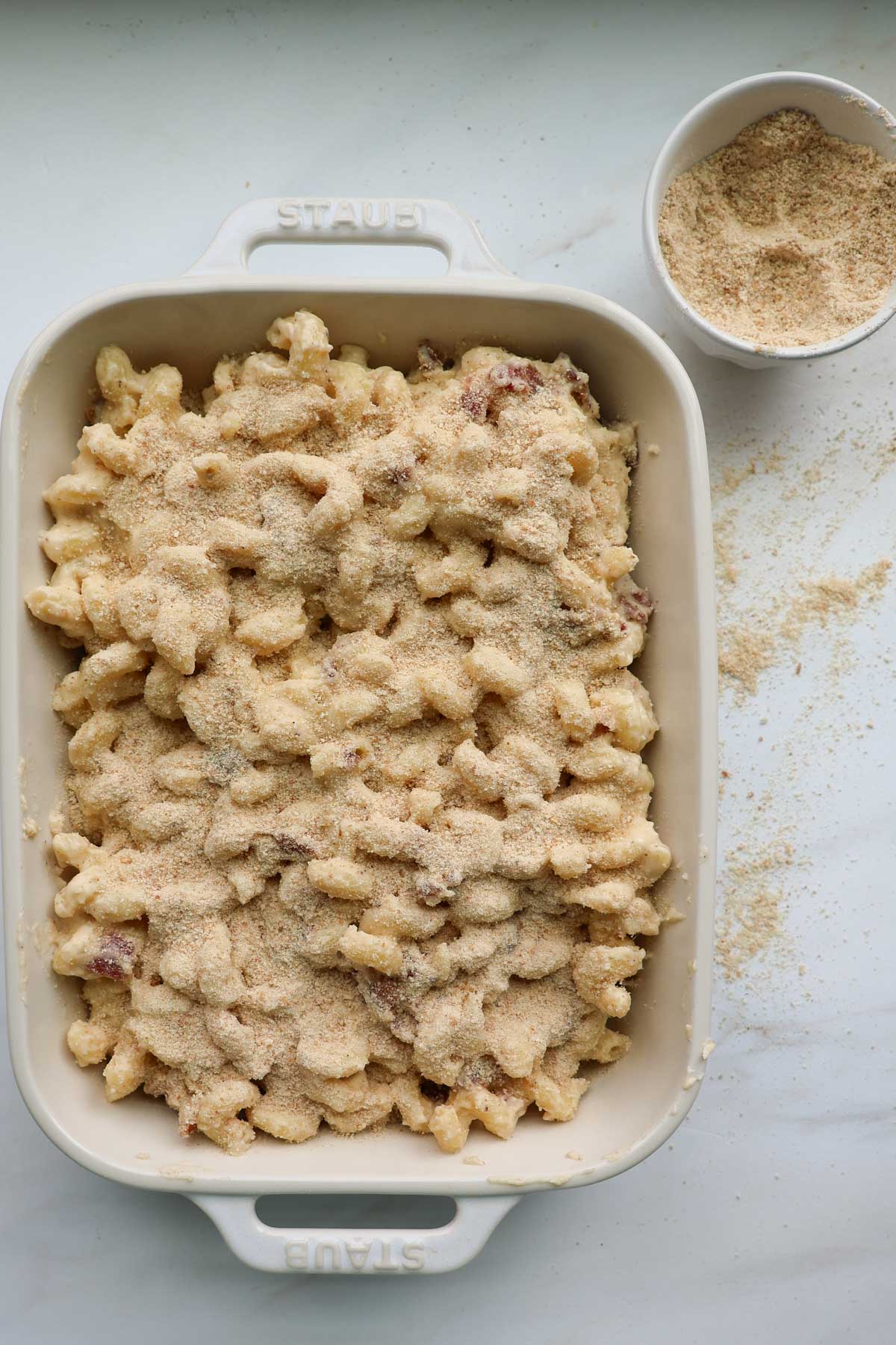 A white, 9 by 13-inch casserole dish filled with bacon mac and cheese, topped with breadcrumbs and a bowl of breadcrumbs nearby.