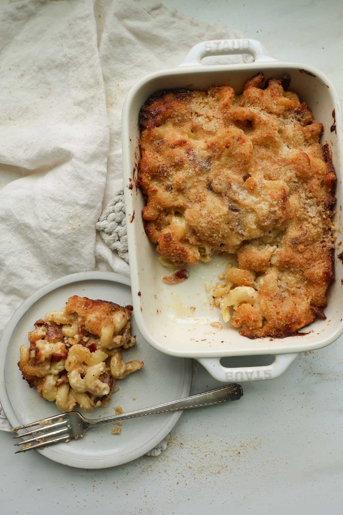 A white, 9 by 13-inch casserole dish filled with baked bacon mac and cheese and a plate with mac and cheese on it.