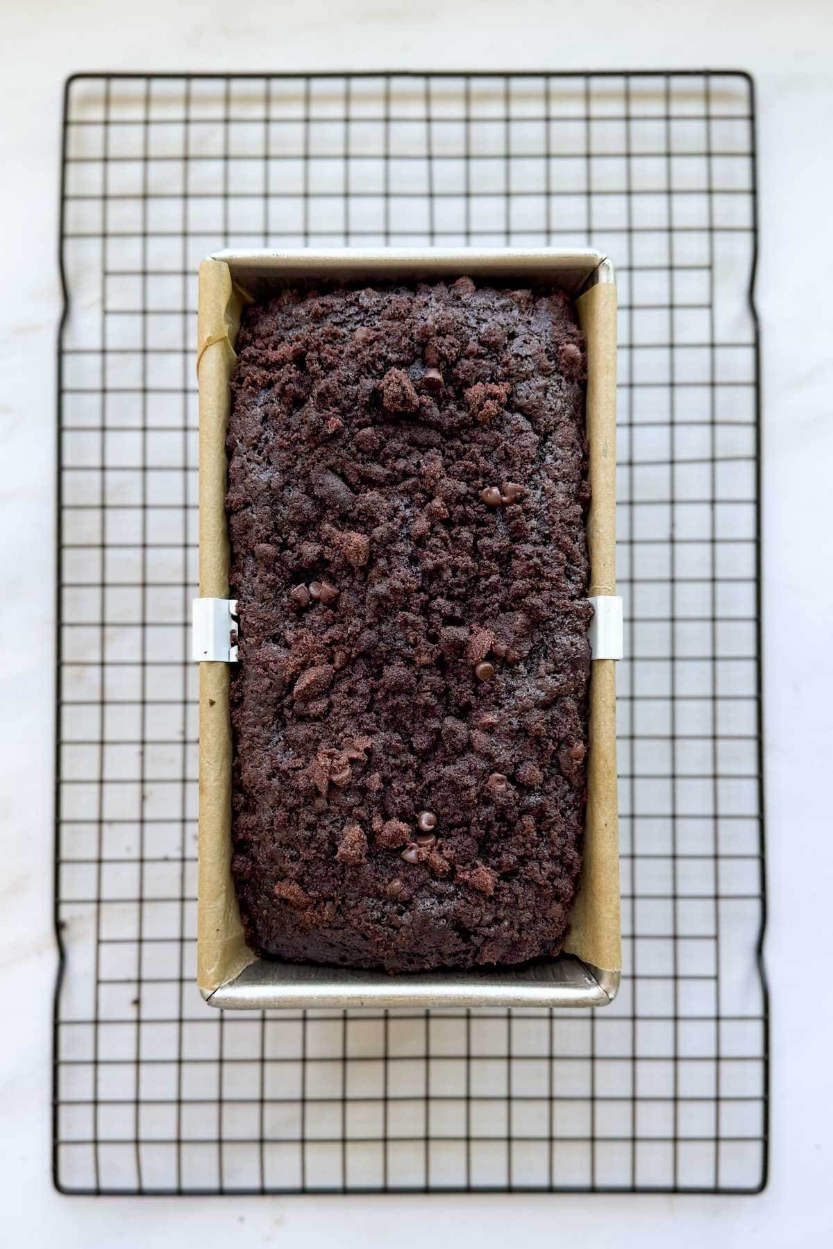 A baked loaf of double chocolate zucchini bread in a loaf pan lined with parchment paper, resing on a wire cooling rack.