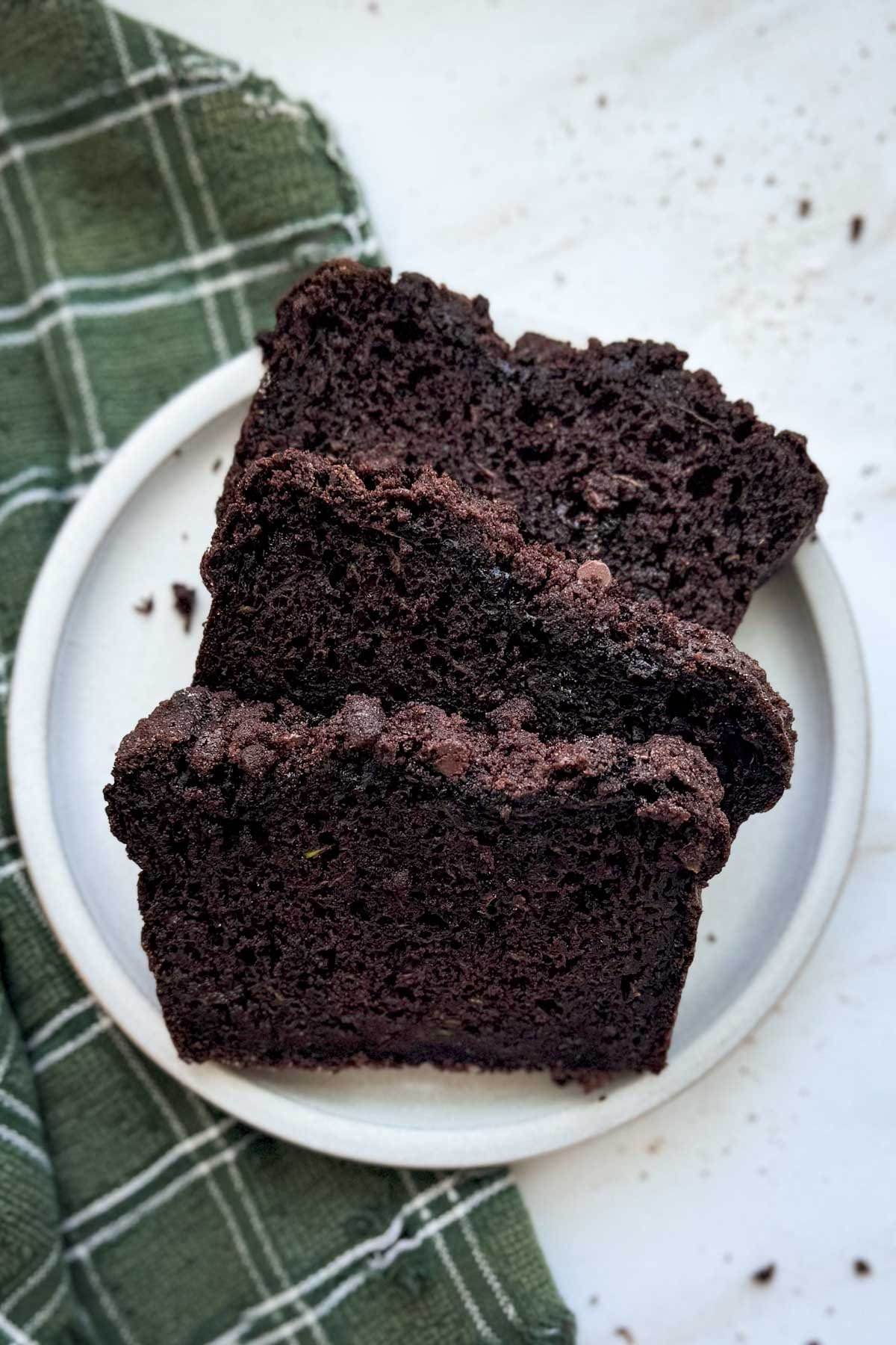 Three slices of double chocolate zucchini bread on a white plate with a green plaid linen underneath it.