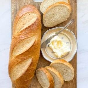 A loaf of french bread on a bread board with sliced french bread and a butter dish.