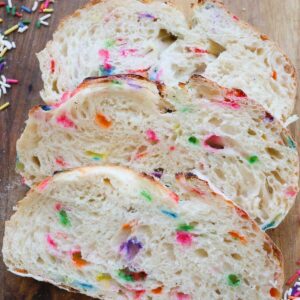 Three slices of funfetti sourdough resting on a wood cutting board with sprinkles around it.
