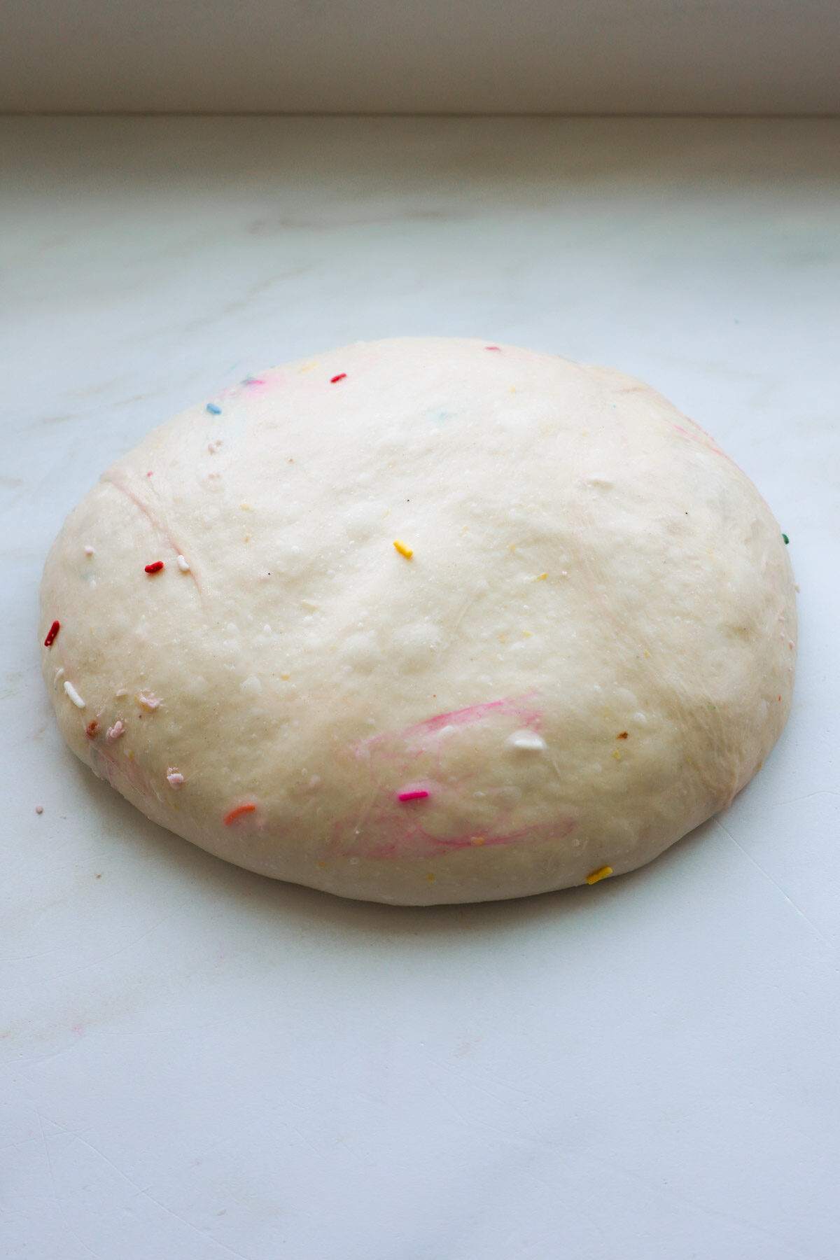 A flattened ball of funfetti sourdough dough resting on a white marble counter.