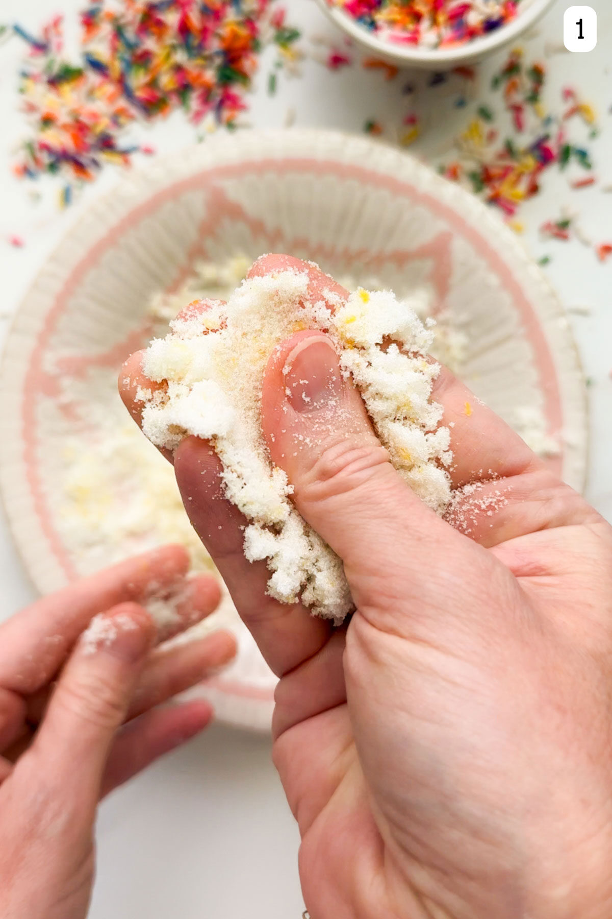 Two hands rubbing sugar and lemon zest together with a bowl and sprinkles on the countertop below.