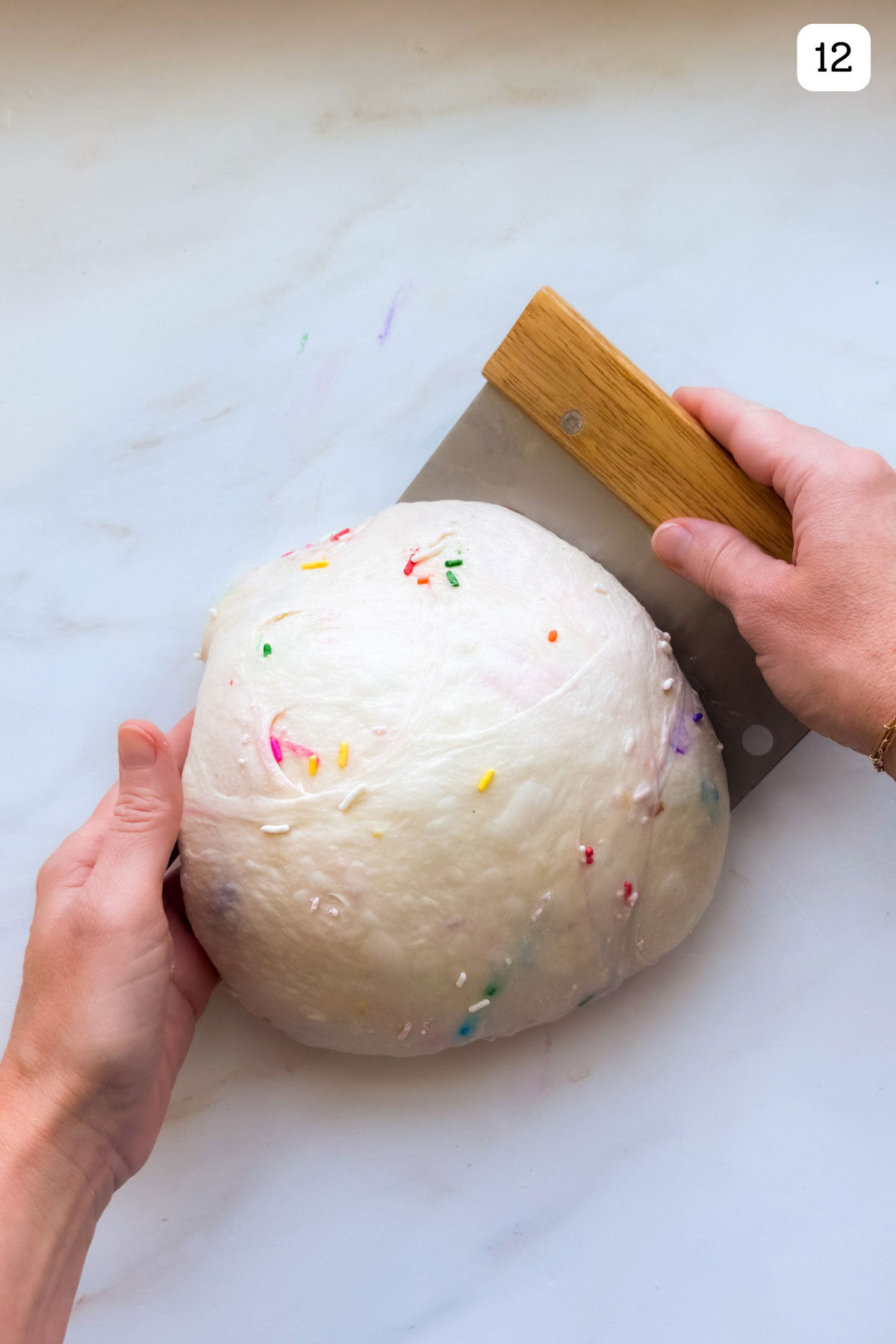 A ball of funfetti sourdough with two hands rounding it out and one hand holding a bench knife.