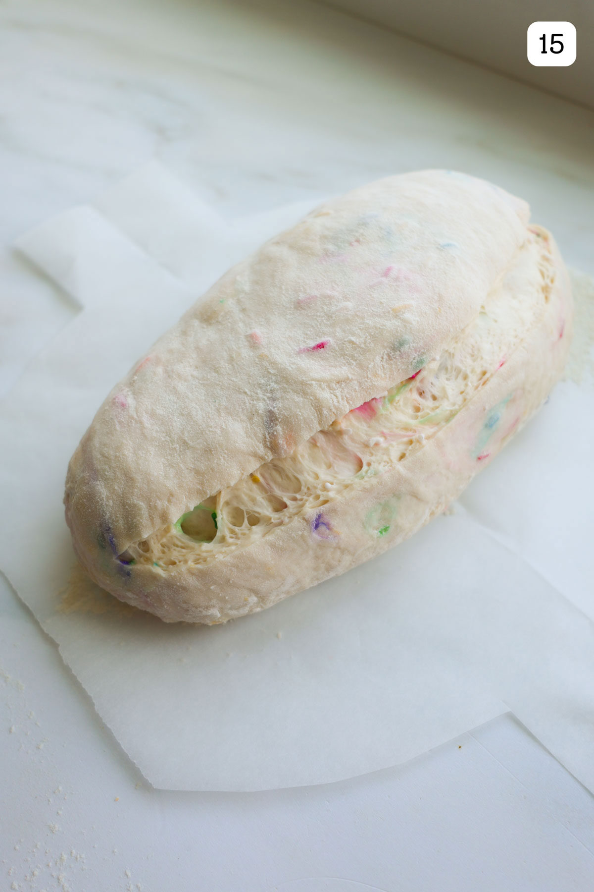 An oval shaped loaf resting on a piece of parchment paper that has a score mark down the the length of the loaf to the right of the center.