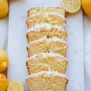 A slice loaf of lemon coconut bread on a white marble cutting board with lemons around it.
