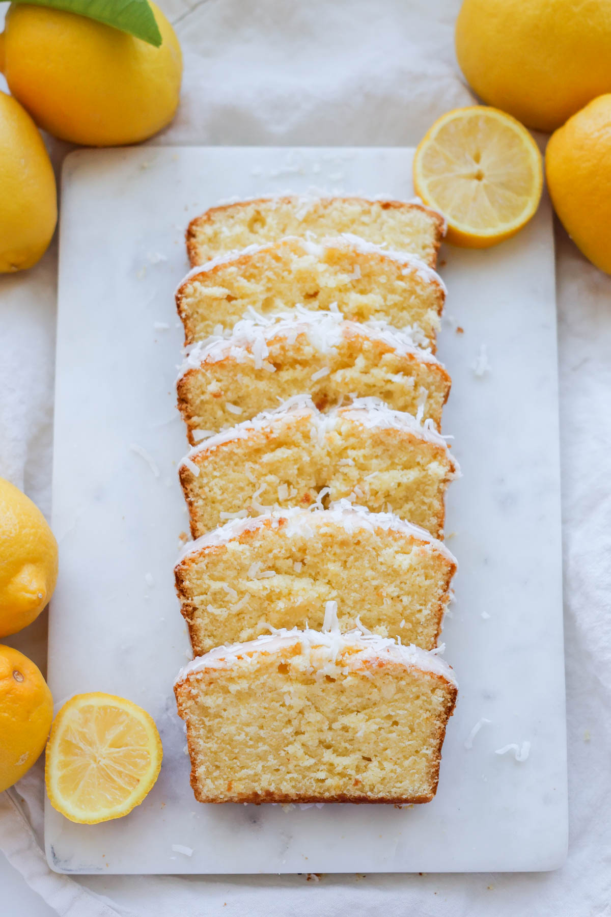 A slice loaf of lemon coconut bread on a white marble cutting board with lemons around it.