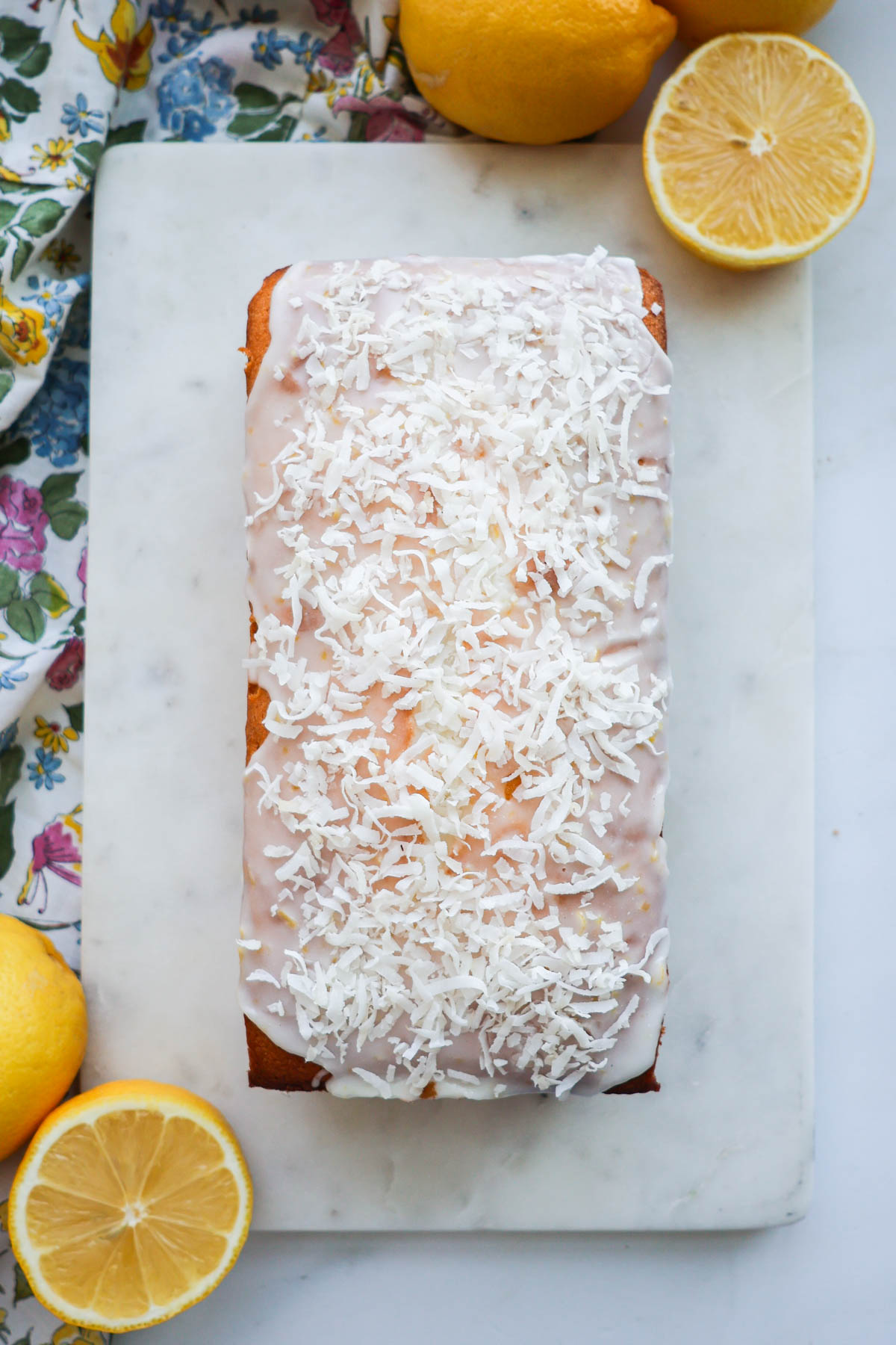 A lemon coconut loaf on a white marble cutting board, surrounded by a floral cloth and lemons.