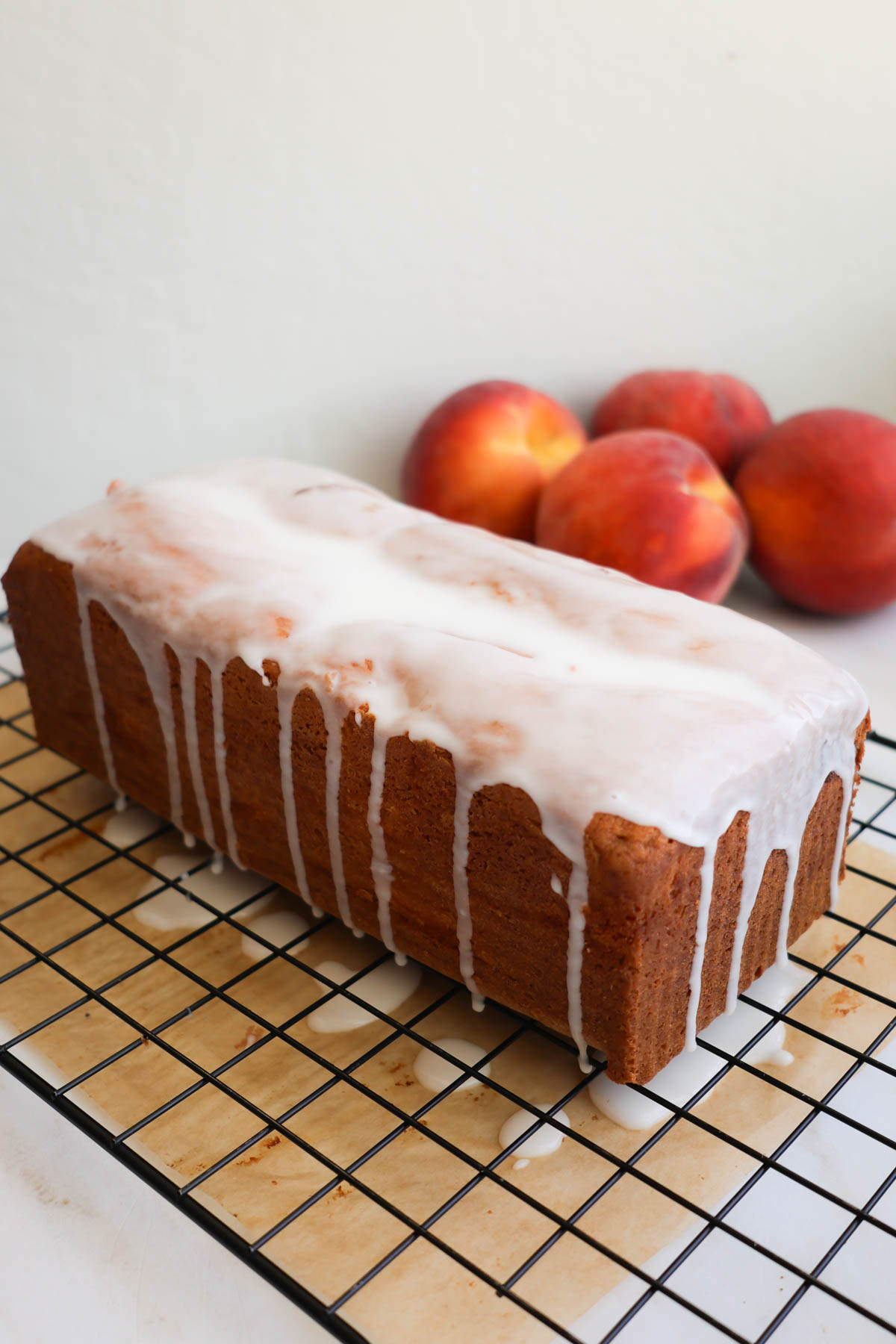 A baked loaf of peach bread with vanilla icing on top, and resting on top of a wire cooling rack with parchment paper underneath.