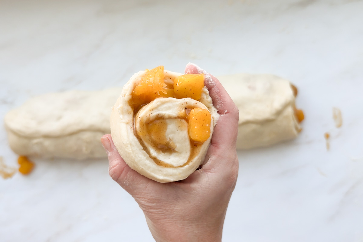 A hand holding an unbaked peach cinnamon roll with dough in the background.