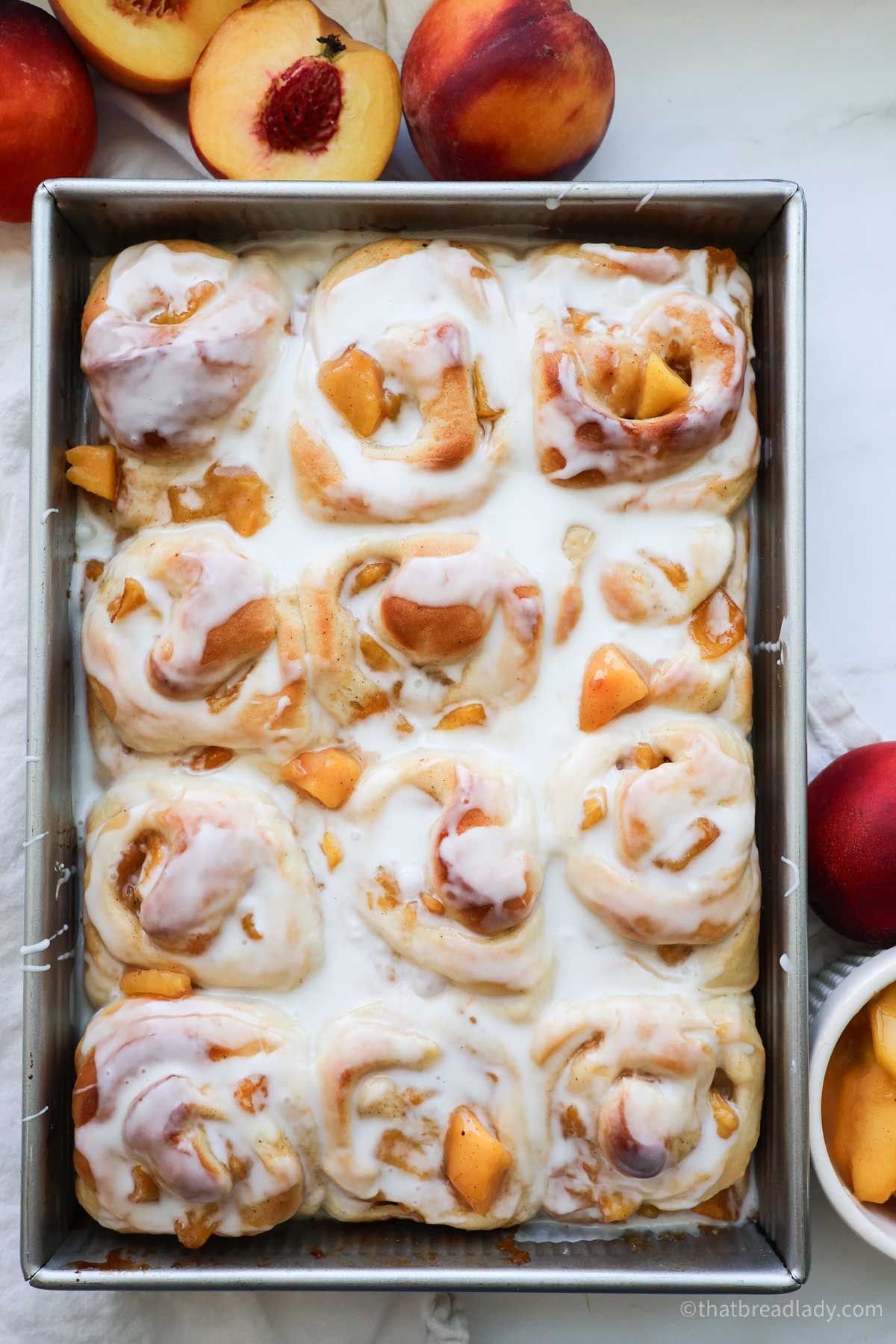 A pan of baked peach cinnamon rolls with icing and peaches all around the pan.