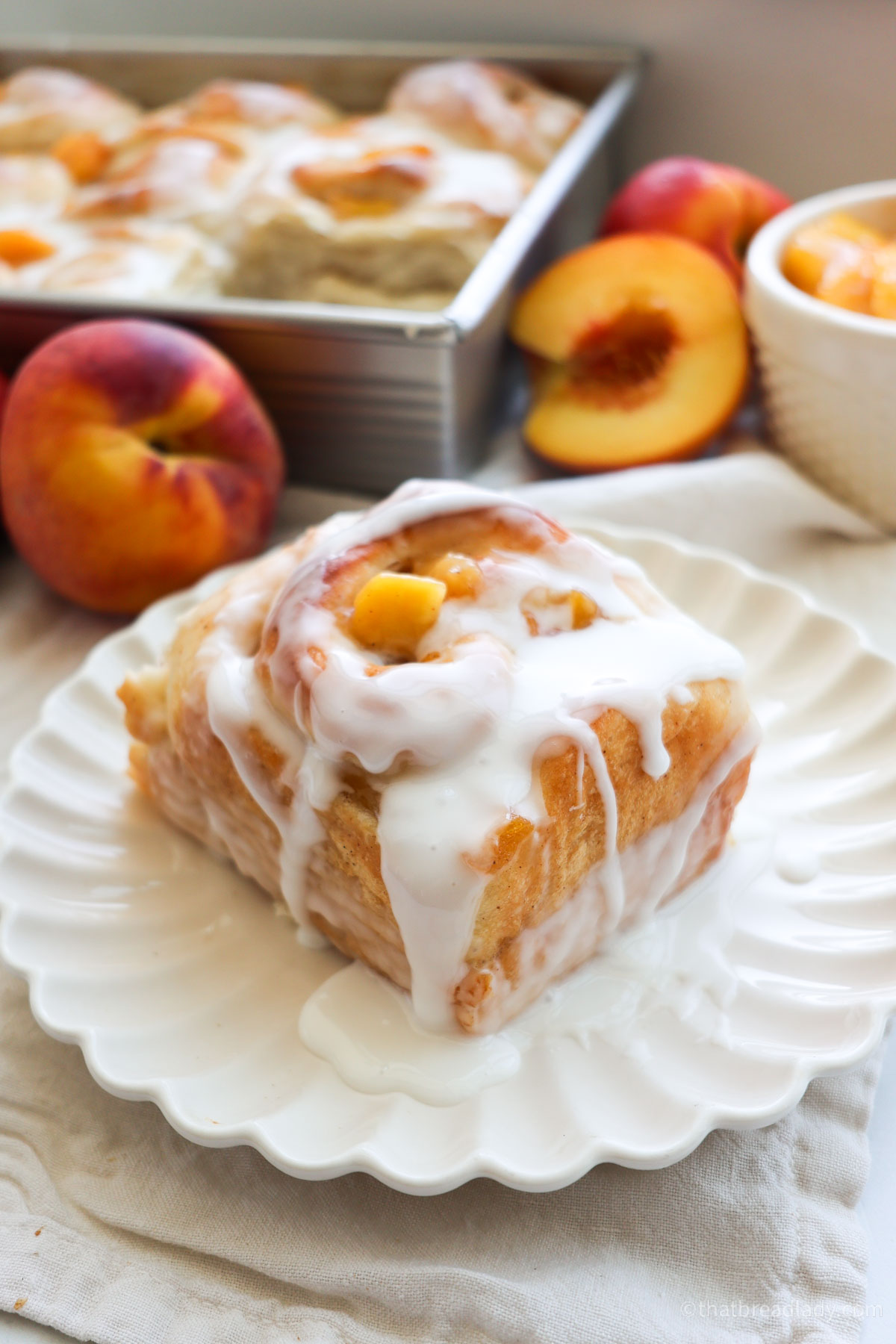 A peach cinnamon roll on a white plate with a pan and peaches in the background.