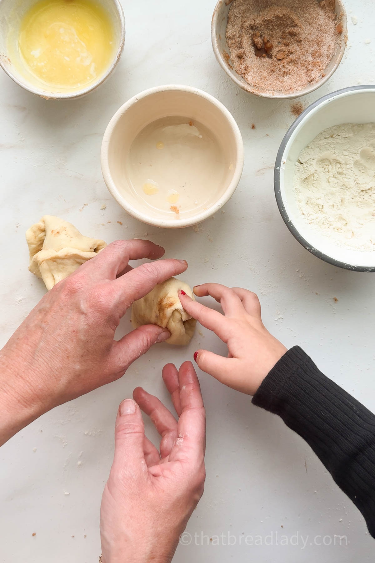 An adult and child's hands shaping dough with bowls of melted butter and cinnamon sugar on the table.