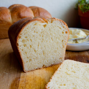 A loaf of sliced brioche on a wood cutting board with butter and more bread in the background.