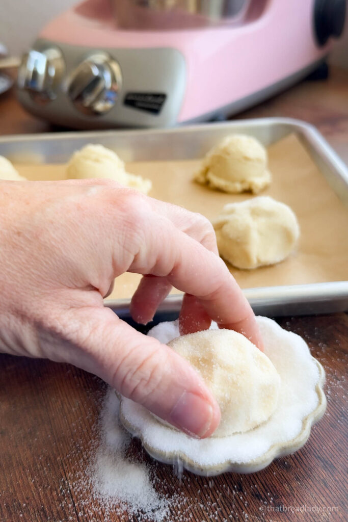 A hand rolling a ball of sugar cookie dough in a small white bowl of sugar.