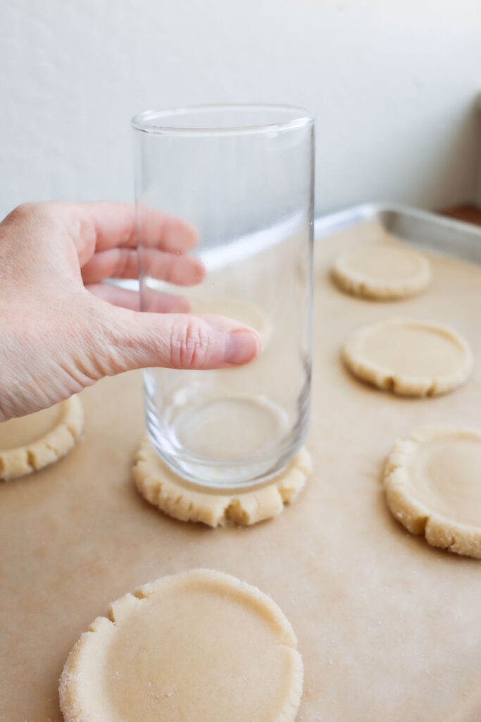 A hand holding a glass, pressing it into a ball of cookie dough, flattening it out.