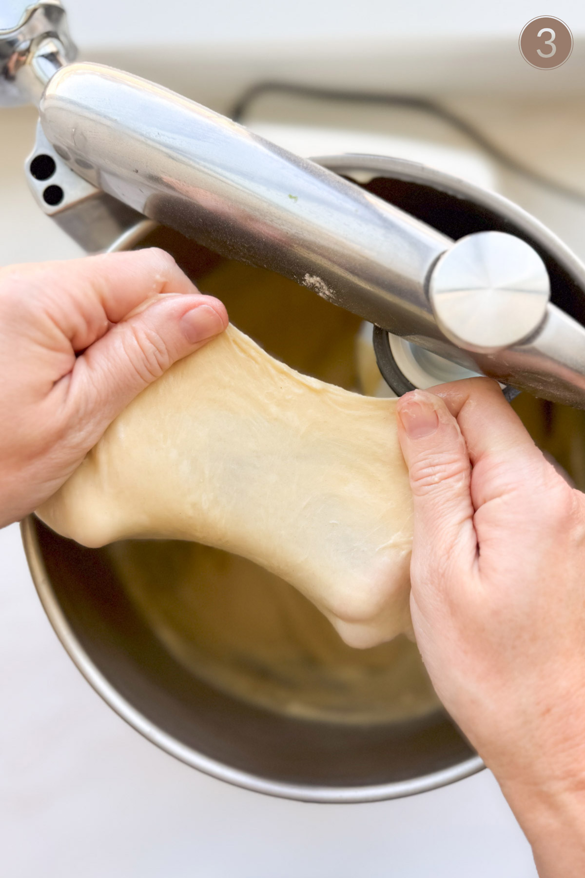 Two hands pulling apart a piece of bread dough, over the bowl of an Ankarsrum stand mixer.