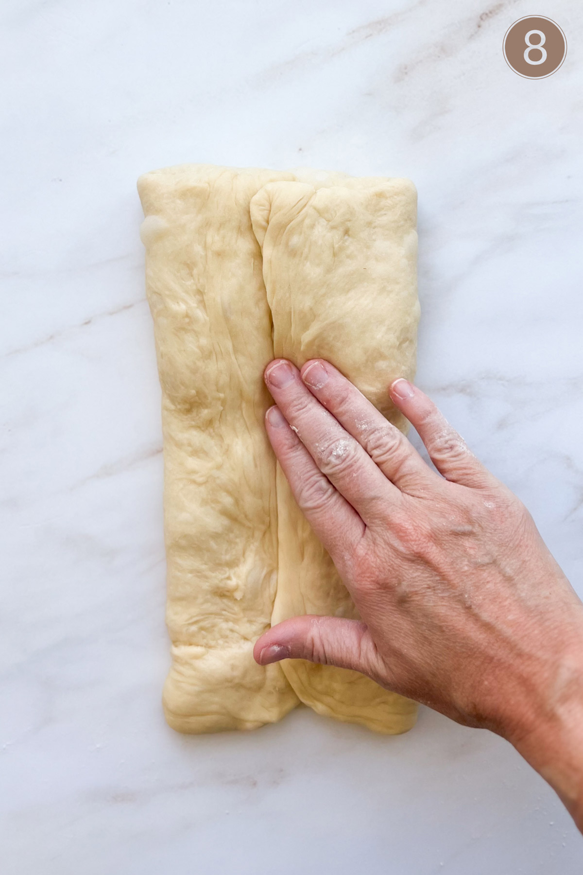 Bread dough folded with each side meeting in the center and a hand pressing down on the center seam.