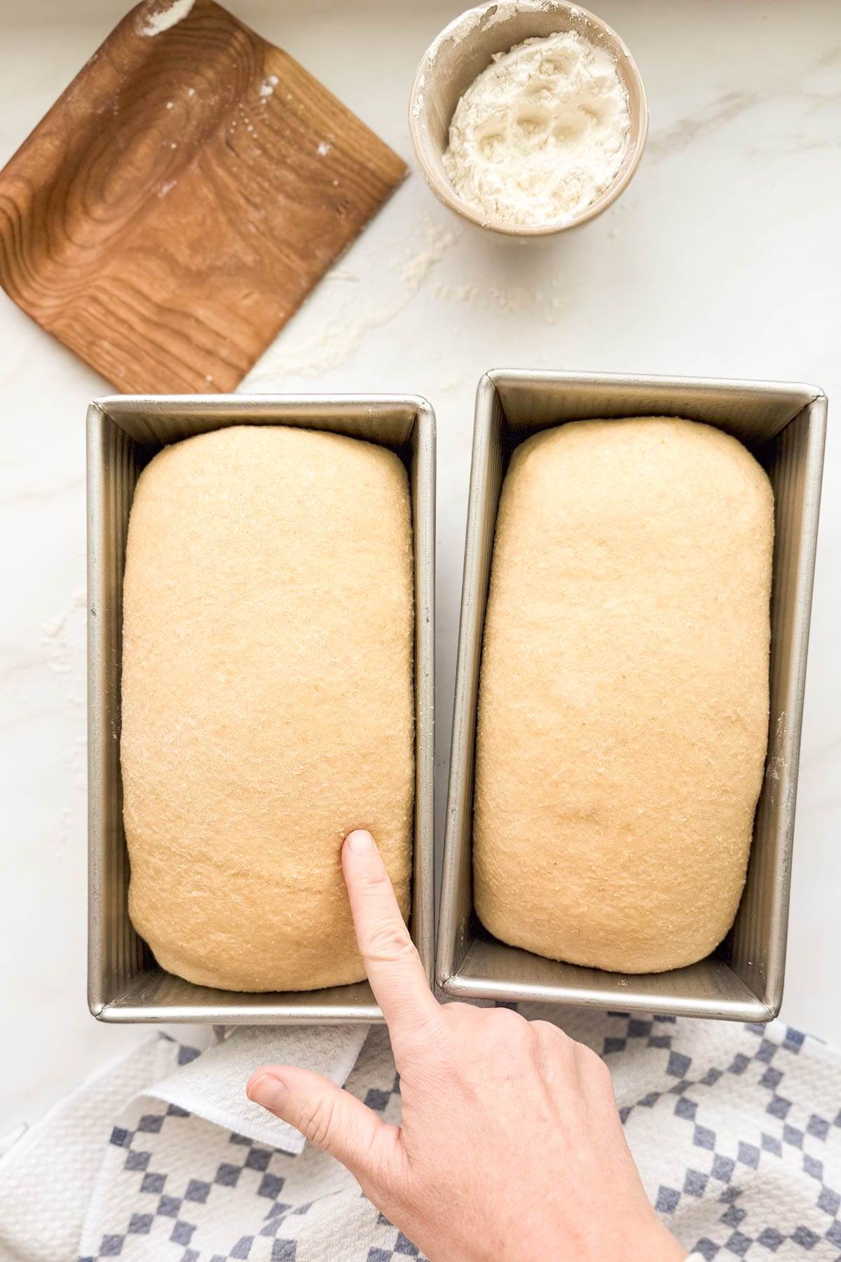 A finger poking the side of one of two loaves of bread dough.